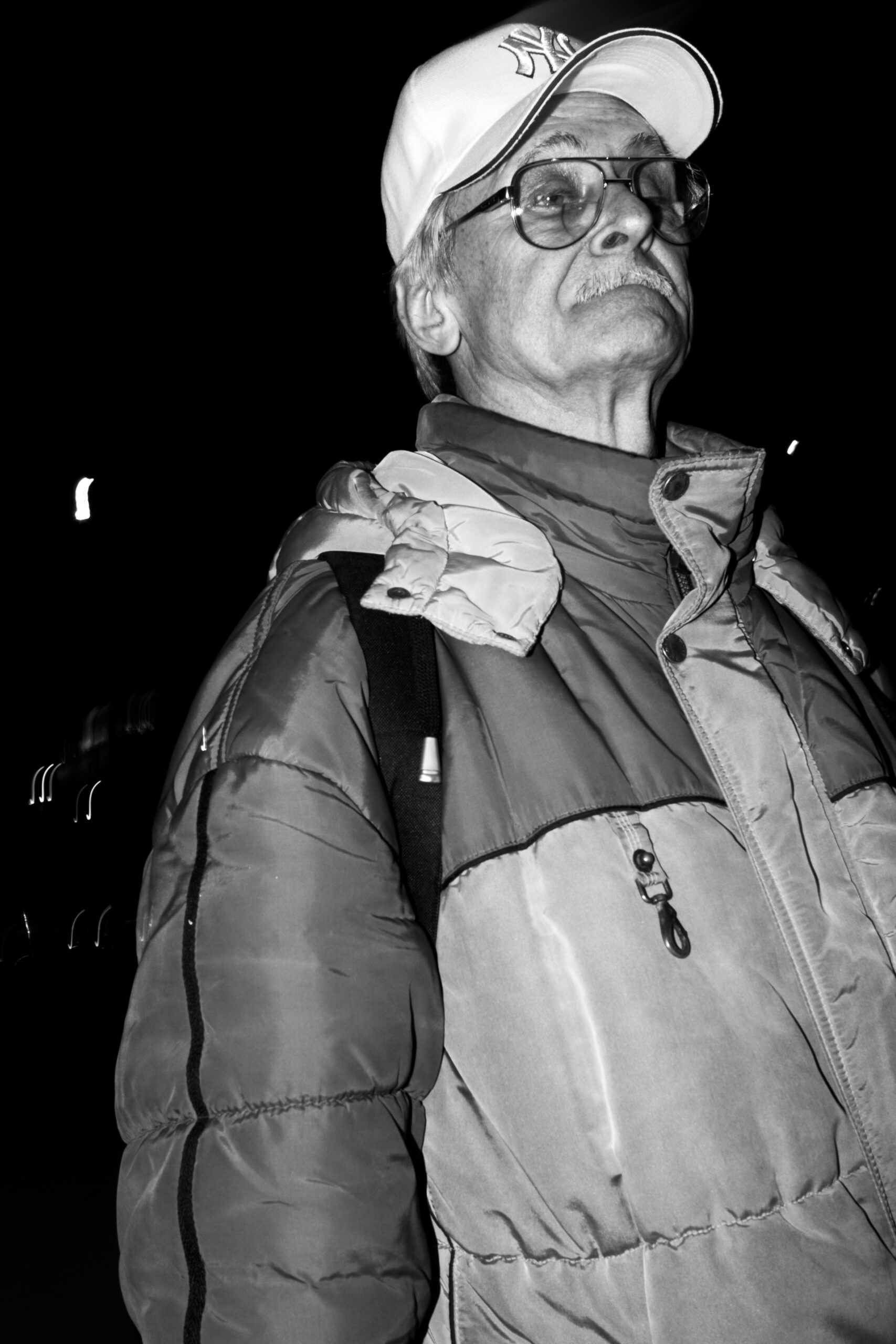 An older man in a cap and padded jacket looking up at night