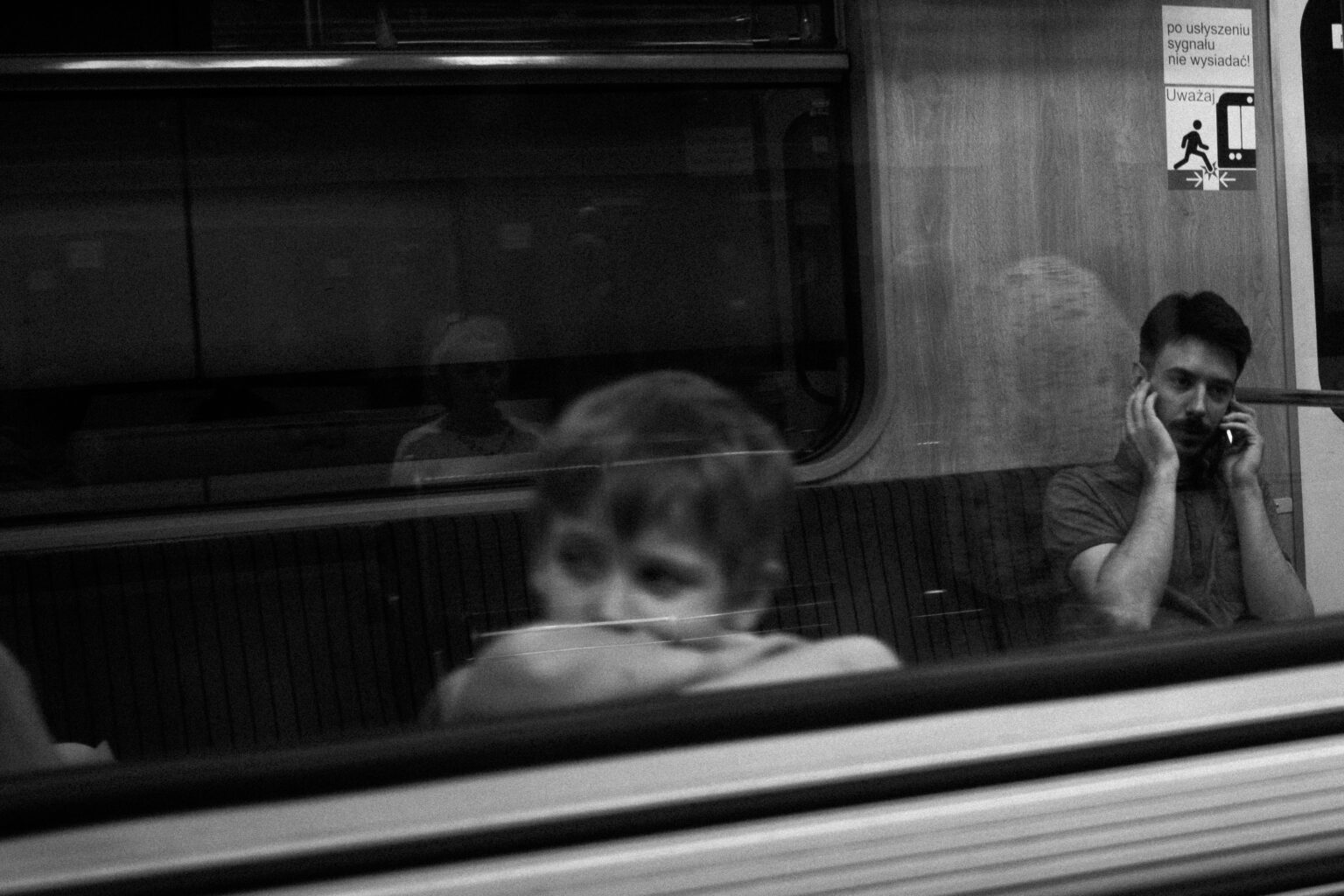 A boy reflected in a metro window while a man talks on the phone
