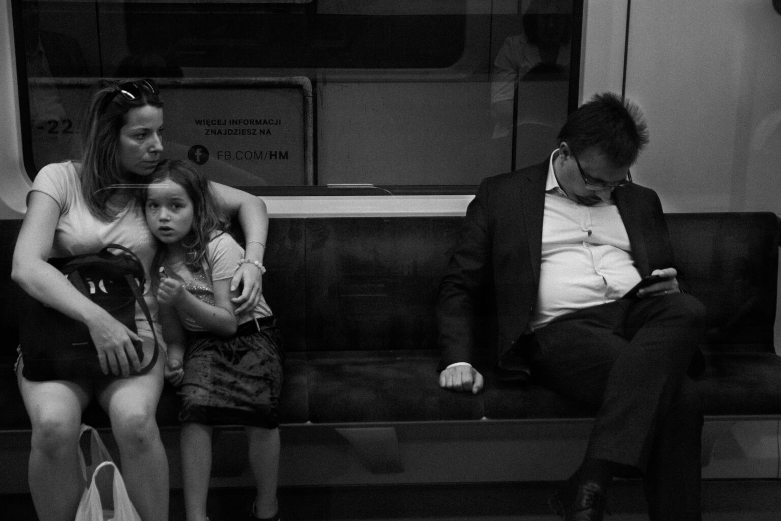 A mother hugging her daughter beside a man reading his phone on a metro seat